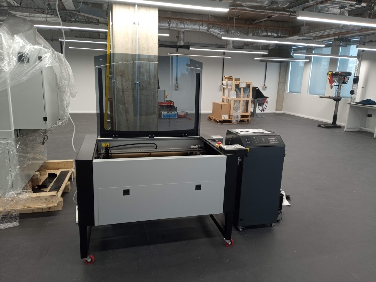 Kent Prestige laser cutter with its lid open beside a fume extraction unit in a workshop